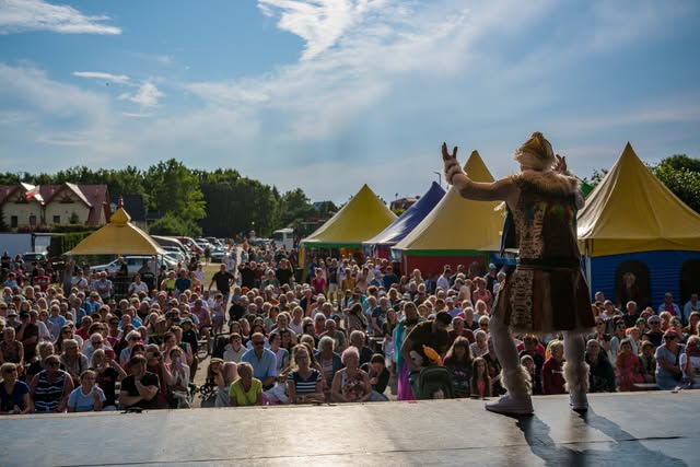 A performer in costume stands at the edge of an outdoor stage, arms raised, facing a crowd of several hundred people under yellow festival tents.