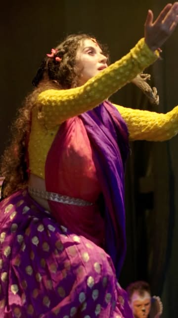 A dancer in a yellow, pink and purple costume reaches upward under stage light.
